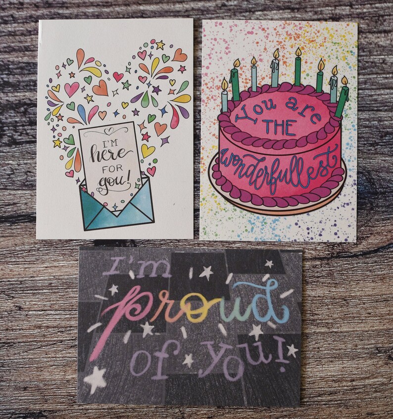 Puede incluir: Tres tarjetas de felicitaci&oacute;n con dise&ntilde;os coloridos. La primera tarjeta tiene una forma de coraz&oacute;n hecha de estrellas y fuegos artificiales coloridos con un sobre y una nota que dice "&iexcl;Estoy aqu&iacute; para ti!" La segunda tarjeta tiene un pastel rosa con velas y el texto "Eres el m&aacute;s maravilloso". La tercera tarjeta tiene un fondo negro con estrellas blancas y el texto "&iexcl;Estoy orgulloso de ti!" en colores arco&iacute;ris.