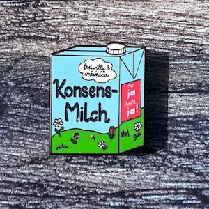 Könnte beinhalten: Ein Anstecker in Form eines Milchkartons mit den Worten "Konsens-Milch" in Blau und Weiß. Der Karton hat einen hellblauen Hintergrund mit einer grünen Basis, Blumen und dem Text "freiwillig & unkenbarbar". Ein rotes Quadrat lautet "nur ja heißt ja!"
