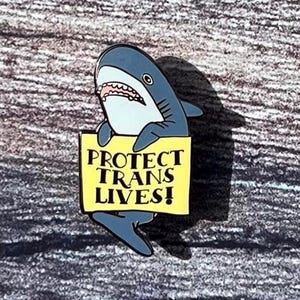 Könnte beinhalten: Ein blauer Hai-Emaille-Pin, der ein gelbes Schild mit der Aufschrift "PROTECT TRANS LIVES!" hält. Der Hai hat einen weißen Bauch, einen weit geöffneten Mund und ein schwarzes Auge. Der Pin befindet sich auf einer strukturierten, grauen Oberfläche.