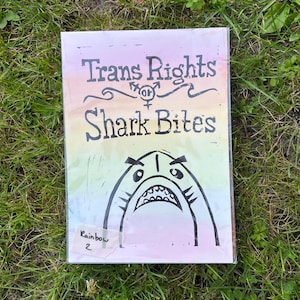 Könnte beinhalten: Ein rechteckiger Druck mit dem Text "Trans Rights" über "Shark Bites" in schwarzer Schrift. Unter dem Text befindet sich ein Cartoon-Hai mit offenem Maul. Der Hintergrund hat einen Regenbogenverlauf. Ein Aufkleber in der unteren linken Ecke lautet "Rainbow 2".