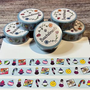 Könnte beinhalten: Fünf Rollen blaues Washi-Tape mit einem weißen Blumenmuster und dem Text "@Baschdlmaidli" auf jeder Rolle. Das Band zeigt ein sich wiederholendes Muster aus Weihnachtskugeln, Zuckerstangen, Schneemännern und Geschenkboxen.