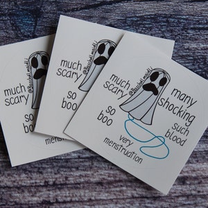 May include: Three white stickers with a ghost illustration and text. The ghost has a sad face and is surrounded by phrases like "much scary" and "so boo." The stickers are on a wooden surface.