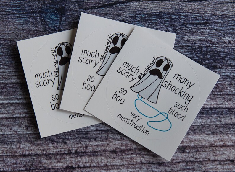 May include: Three white stickers with a ghost illustration and text. The ghost has a sad face and is surrounded by phrases like "much scary" and "so boo." The stickers are on a wooden surface.