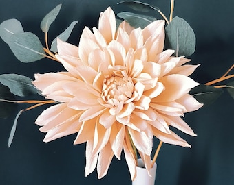 Op de afbeelding: Een enkele, grote, perzikkleurige papieren bloem in een witte vaas met groene papieren bladeren. De vaas staat op een lichtbruin houten oppervlak.