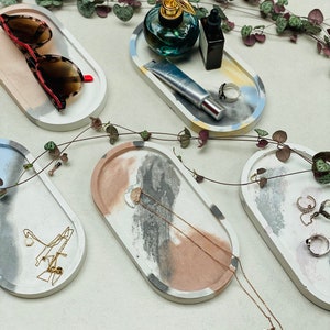 May include: Four oval concrete trays with abstract designs in shades of gray, blue, pink, and yellow. The trays are filled with various items, including sunglasses, a perfume bottle, a tube of lotion, a necklace, and rings.