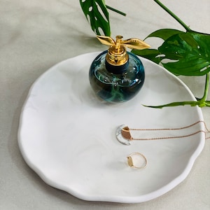 May include: A white ceramic tray with a wavy edge, featuring a blue glass perfume bottle with a gold top, a gold ring, and a gold necklace with a white and gold pendant.