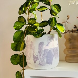 May include: A white ceramic plant pot with a lavender abstract design, holding a trailing plant with green and yellow leaves. The pot sits on a white shelf, showcasing a modern, minimalist aesthetic.