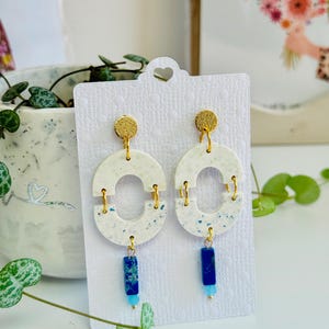 May include: A pair of handmade earrings featuring a white, speckled, oval design with gold-colored accents. The earrings have a blue and gold bead at the bottom. The earrings are displayed on a white card.