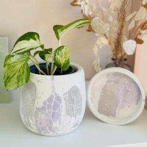 Puede incluir: Un macetero de cer&aacute;mica blanca con un dise&ntilde;o marmoleado de color p&uacute;rpura claro, que presenta una planta verde y blanca variegada. El macetero tiene un platillo a juego.