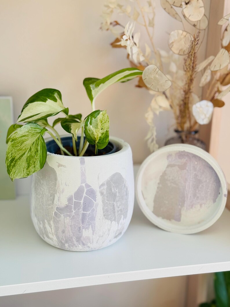 Puede incluir: Un macetero de cer&aacute;mica blanca con un dise&ntilde;o marmoleado de color p&uacute;rpura claro, que presenta una planta verde y blanca variegada. El macetero tiene un platillo a juego.