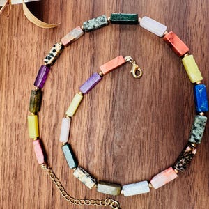 Puede incluir: Un collar colorido hecho de cuentas rectangulares de piedras preciosas. Las cuentas son de varios tonos de verde, azul, morado, rosa y amarillo. El collar tiene un cierre dorado.