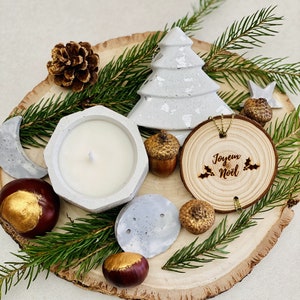 Peut inclure: Une composition festive avec une bougie blanche dans un support en béton géométrique, un sapin de Noël en béton et une décoration en bois avec le texte "Joyeux Noël". L'ensemble comprend des glands, des châtaignes et des aiguilles de pin sur une tranche de bois.