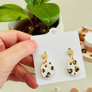 Peut inclure: Boucles d'oreilles en forme de goutte d'eau, crème avec un imprimé léopard noir et beige. Elles sont ornées d'un accent doré scintillant. Les boucles d'oreilles sont présentées sur une carte blanche.