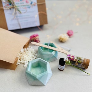 May include: A DIY candle making kit with two teal green concrete candle holders, a small bottle of essential oil, a bag of white wax flakes, a wooden wick, and a glass vial filled with dried flowers.