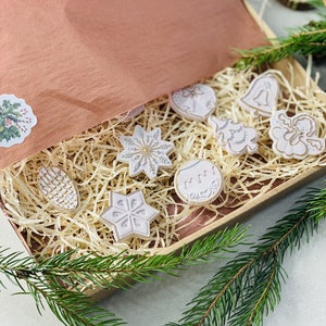 Peut inclure: Une boîte cadeau en carton remplie de copeaux de bois et d'ornements en céramique de couleur claire, incluant une étoile, une cloche, un ange, une pomme de pin et un sapin. La boîte est doublée de papier de soie cuivré et porte un autocollant houx.