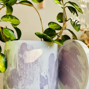 May include: A white and lavender marbled concrete planter with a matching lid. The planter is round and holds a green plant with variegated leaves. The lid is round and rests beside the planter. The background is a neutral color.