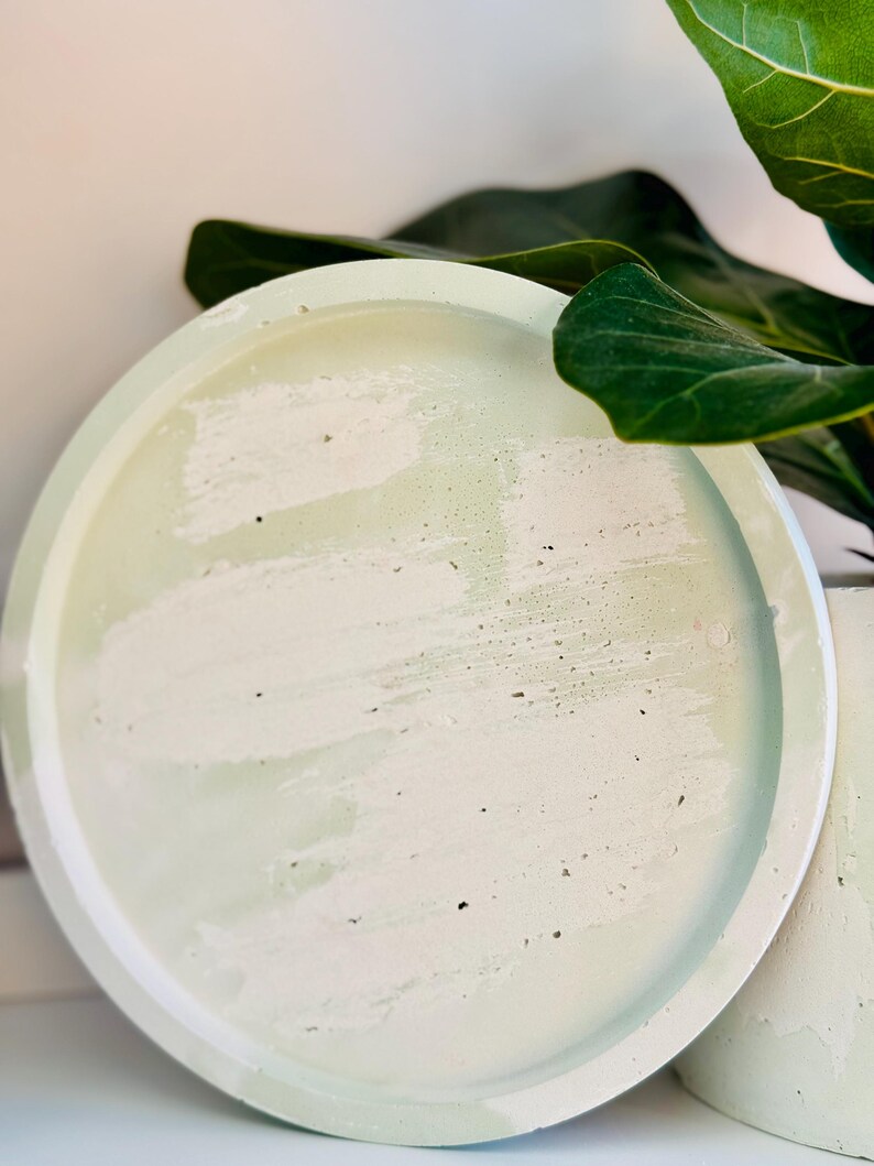 May include: A round, light green concrete tray with a white and green mottled surface. The tray has a raised edge and is next to a matching concrete cylinder. A large green plant leaf is in the background.