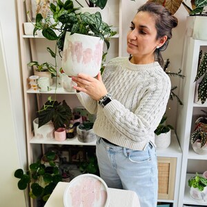 May include: A white ceramic planter with pink paint strokes, filled with green leafy plants. A matching tray sits on a table in front of a shelf with other plants and planters. The planter is approximately 8 inches in diameter.