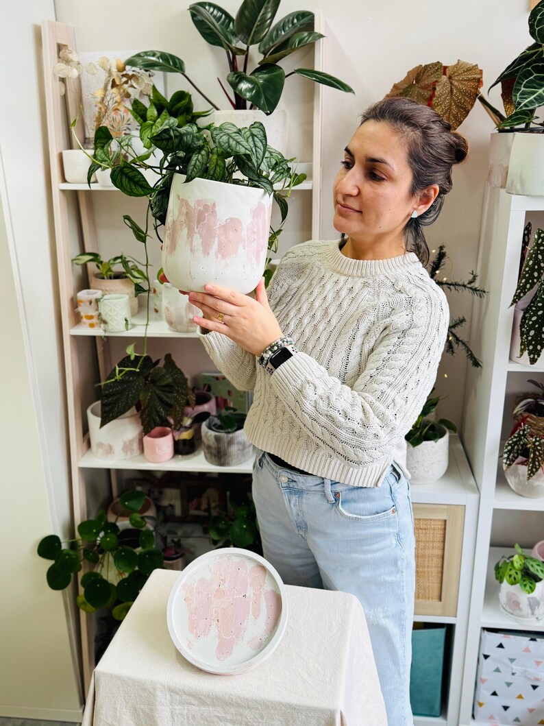 May include: A white ceramic planter with pink paint strokes, filled with green leafy plants. A matching tray sits on a table in front of a shelf with other plants and planters. The planter is approximately 8 inches in diameter.