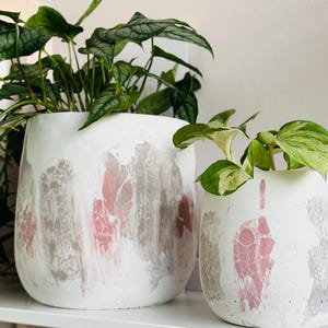 May include: Two white ceramic plant pots with pink and gray abstract designs. The pots are filled with green leafy plants and sit on a white shelf. The pots have a textured, painted appearance.