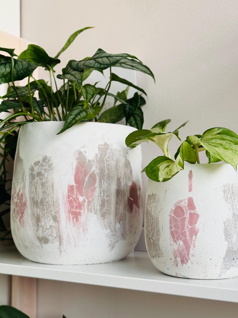 May include: Two white ceramic plant pots with pink and gray abstract designs. The pots are filled with green leafy plants and sit on a white shelf. The pots have a textured, painted appearance.