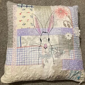 May include: Square patchwork cushion with a hand-drawn bunny design. The bunny has pink-lined ears and a pink nose. The cushion incorporates various fabric patterns, including floral, gingham, and script. A white lace flower is attached.