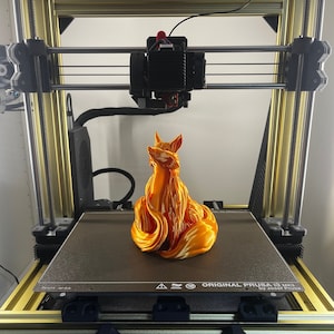 Puede incluir: Una impresora 3D con marco dorado está imprimiendo una figura de zorro naranja y blanco. El panel de control de la impresora muestra información de temperatura y progreso. La impresora está etiquetada como "ORIGINAL PRUSA I3 MK3S+".