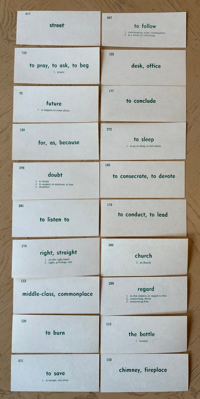Op de afbeelding: Een set van 16 witte indexkaarten met groene tekst. Elke kaart heeft een nummer en een woord of zin met een definitie. De woorden en zinnen omvatten "straat", "bidden, vragen, smeken", "toekomst", "want, zoals, omdat", "twijfel", "luisteren", "recht, recht", "middenklasse, alledaags", "branden", "redden", "volgen", "bureau, kantoor", "concluderen", "slapen", "wijden, wijden", "leiden, leiden", "kerk", "aandacht", "de fles", "schoorsteen, open haard".