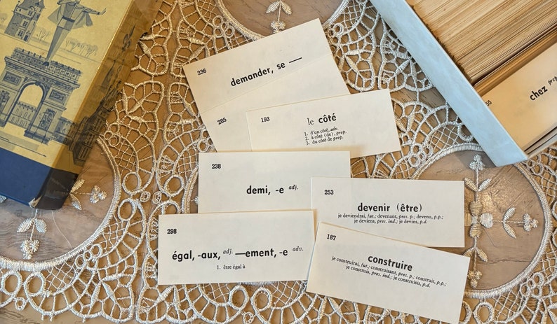 Op de afbeelding: Close-up van verschillende witte indexkaarten met Franse woorden en definities in zwarte inkt. De kaarten zijn gerangschikt op een wit kant kussen. De woorden omvatten "demander", "le c&ocirc;t&eacute;", "demi", "&eacute;gal", "devenir" en "construire".