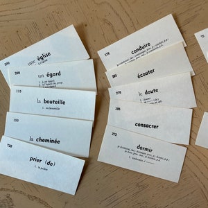 Op de afbeelding: Een set witte indexkaarten met Franse woordenschatwoorden geschreven in zwarte inkt. De kaarten zijn in een waaier op een lichtbruin houten oppervlak gerangschikt.