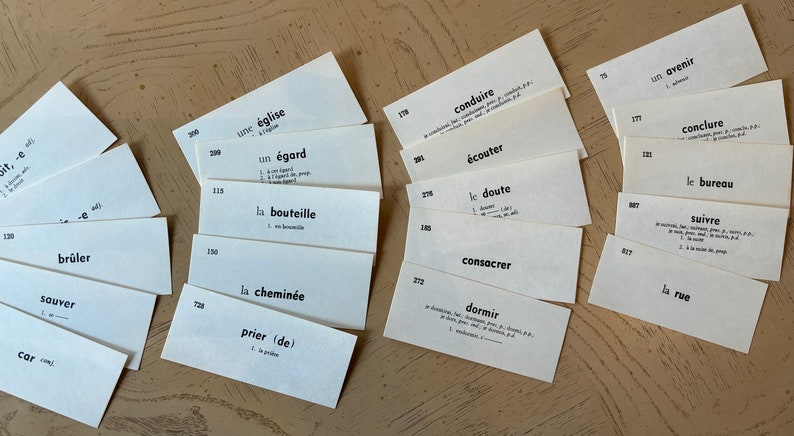 Op de afbeelding: Een set witte indexkaarten met Franse woordenschatwoorden geschreven in zwarte inkt. De kaarten zijn in een waaier op een lichtbruin houten oppervlak gerangschikt.