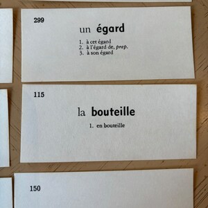 Op de afbeelding: Drie witte indexkaarten met zwarte tekst. De eerste kaart heeft het nummer 299 en het woord "un &eacute;gard" met drie definities. De tweede kaart heeft het nummer 115 en het woord "la bouteille" met &eacute;&eacute;n definitie. De derde kaart heeft het nummer 150 en het woord "la chemin&eacute;e".
