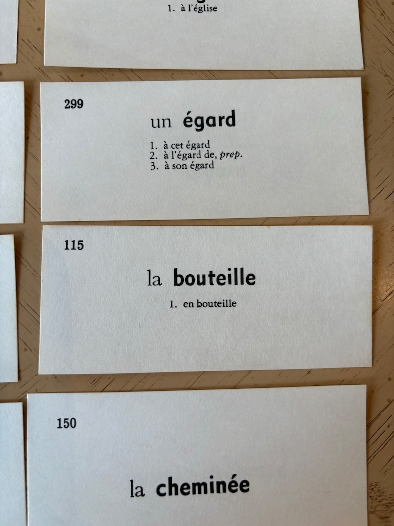 Op de afbeelding: Drie witte indexkaarten met zwarte tekst. De eerste kaart heeft het nummer 299 en het woord "un &eacute;gard" met drie definities. De tweede kaart heeft het nummer 115 en het woord "la bouteille" met &eacute;&eacute;n definitie. De derde kaart heeft het nummer 150 en het woord "la chemin&eacute;e".