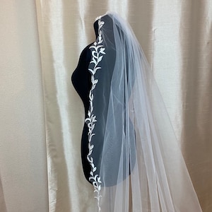 May include: A white bridal veil with a delicate floral pattern along the edge. The veil is made of soft tulle and is perfect for a wedding ceremony.