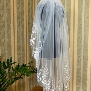 May include: A white bridal veil with delicate floral embroidery along the edge. The veil is made of soft tulle and is designed to be worn over the head.