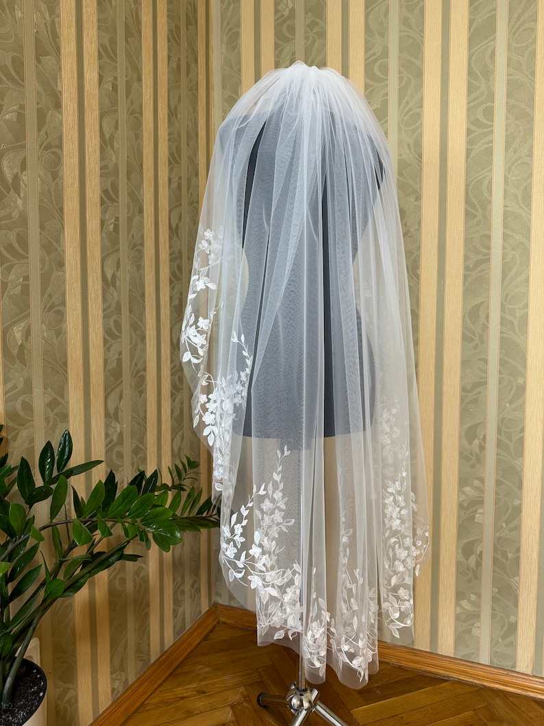 May include: A white bridal veil with delicate floral embroidery along the edge. The veil is made of soft tulle and is designed to be worn over the head.