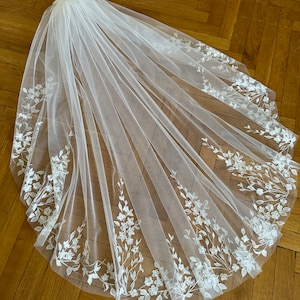 May include: A white bridal veil with delicate floral embroidery along the edges. The veil is made of soft tulle and has a classic design.