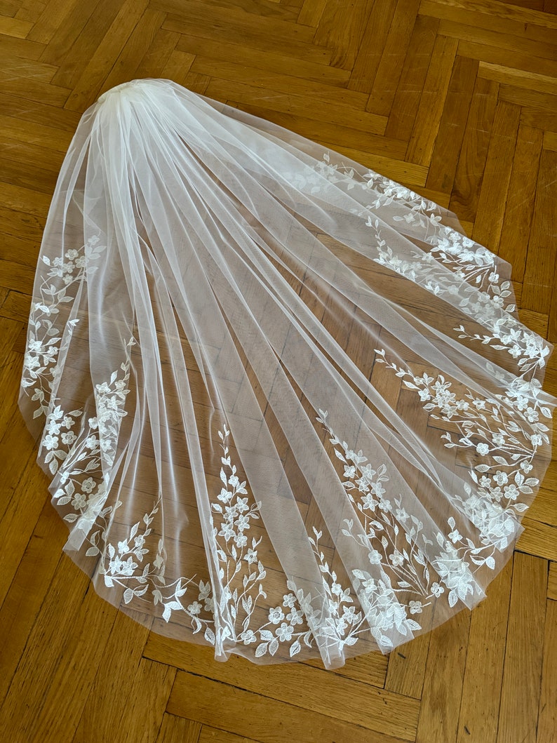 May include: A white bridal veil with delicate floral embroidery along the edges. The veil is made of soft tulle and has a classic design.