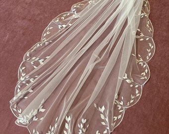 May include: A sheer, ivory-colored bridal veil with a scalloped edge. The veil is adorned with delicate, embroidered leaf patterns along the border. The veil is made of a lightweight fabric, creating a soft, flowing appearance.
