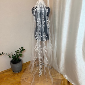 Peut inclure: Un voile de mariée blanc transparent orné de motifs de feuilles brodées. Le voile est présenté sur un mannequin noir, mettant en valeur sa longueur et son design. L'arrière-plan est composé d'un mur neutre et d'un rideau blanc.