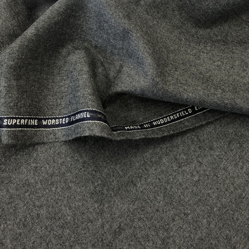Op de afbeelding: Close-up van een grijze stof met een label met de tekst "Superfine Worsted Flannel Made in Huddersfield England".