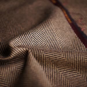 Op de afbeelding: Close-up van een bruine en zwarte herringbone stof met een rood en gouden geweven label.
