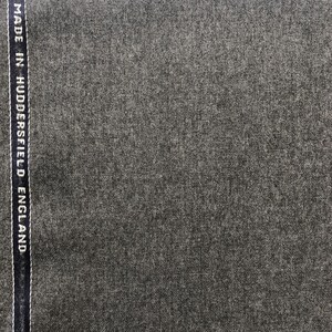 Op de afbeelding: Close-up van een donkergrijze stof met een subtiele textuur. De stof heeft een label met de tekst "MADE IN HUDDERSFIELD ENGLAND".