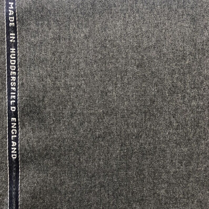 Op de afbeelding: Close-up van een donkergrijze stof met een subtiele textuur. De stof heeft een label met de tekst "MADE IN HUDDERSFIELD ENGLAND".