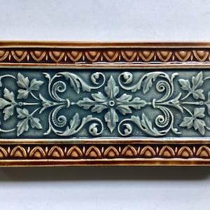 May include: Rectangular ceramic tile featuring a detailed floral and scroll design in blue-green and cream. The tile is bordered by a brown and tan decorative pattern. A classic decorative element.