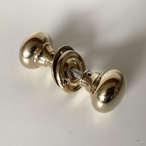 May include: A pair of polished brass door knobs. The knobs are oval and connected by a metal rod. The door knobs are mounted on a circular backplate. The brass has a shiny, reflective surface.
