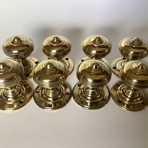 May include: Eight polished brass door knobs. Each knob has a rounded top with concentric rings and a circular base. The knobs are arranged in two rows of four, displaying their reflective, golden finish. The design is classic and timeless.