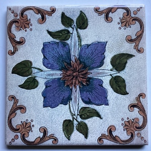 May include: A vintage ceramic tile featuring a floral design. The tile depicts a large blue flower with a brown centre, surrounded by green leaves and gold scrollwork.