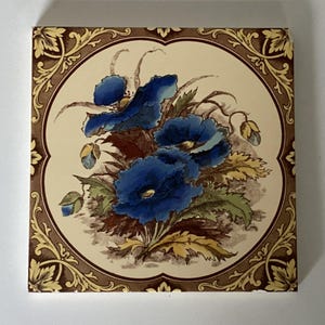 May include: Square ceramic tile with a floral design. The central image displays vivid blue flowers with green and yellow leaves on a cream background. The tile is framed by a brown border with ornate, gold-coloured floral patterns.