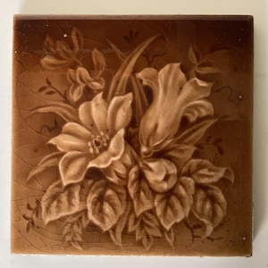 May include: Square ceramic tile featuring a floral design in shades of brown. The flowers and leaves are detailed with a lighter brown, contrasting against the darker background. The tile has a cracked, aged appearance.